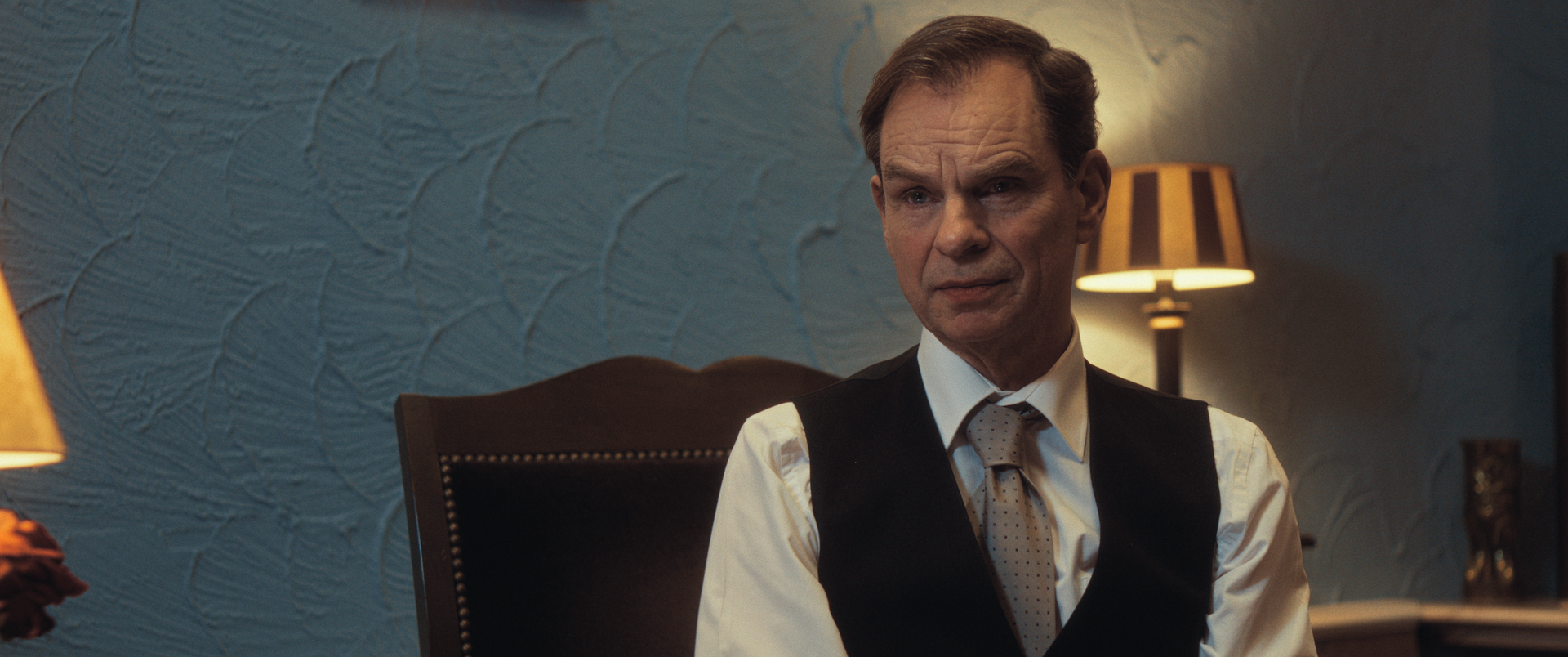 Een man, gekleed in een wit overhemd en een donker vest, zit in een kantoor met blauwe muren en twee lampen en kijkt naar de camera.