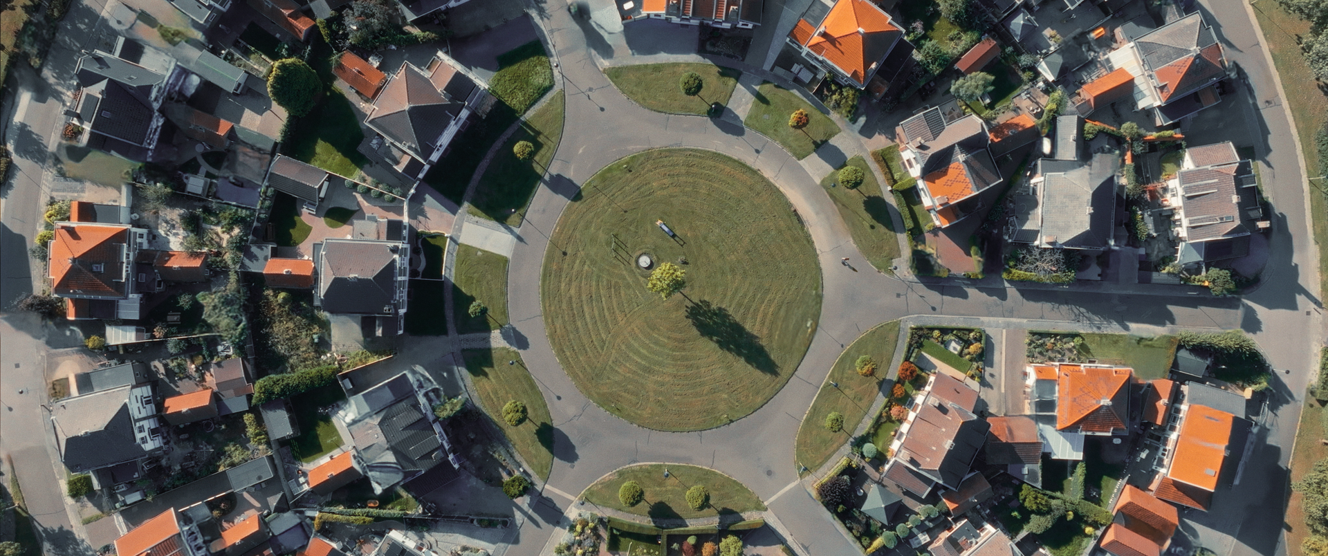 Een luchtfoto van een cirkelvormige woonwijk met een centrale, met gras begroeide rotonde, omringd door huizen.