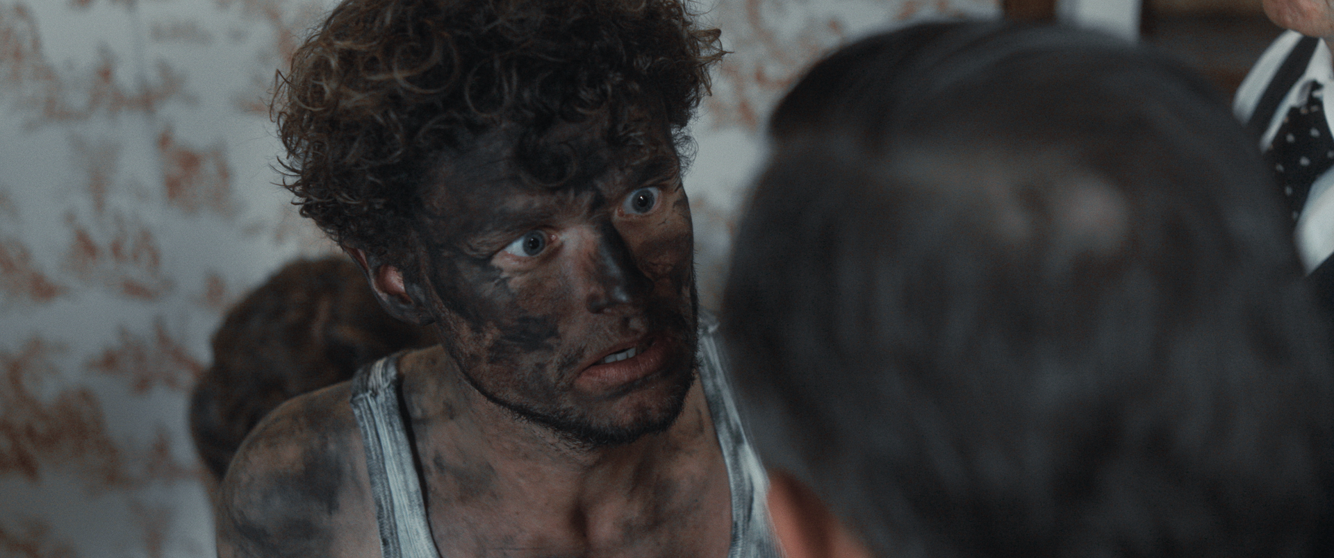 Iemand met een met roet bedekt gezicht kijkt intens naar een ander, met een uitdrukking van verbazing of schok.