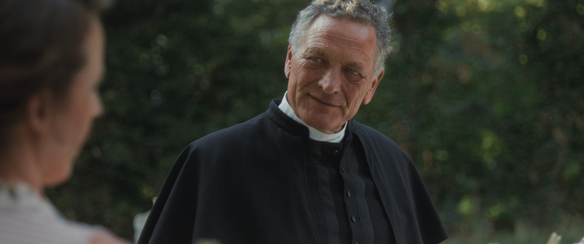 Een man in een zwart geestelijk overhemd met kraag kijkt naar een persoon buiten beeld in een groene buitenomgeving.