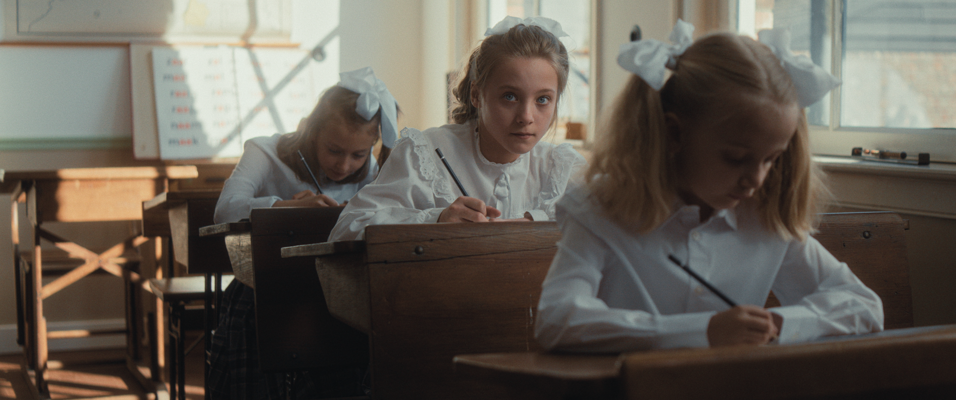 Drie leerlingen in witte shirts en met strikjes in hun haar zitten aan houten bureaus te schrijven in een zonovergoten klaslokaal.