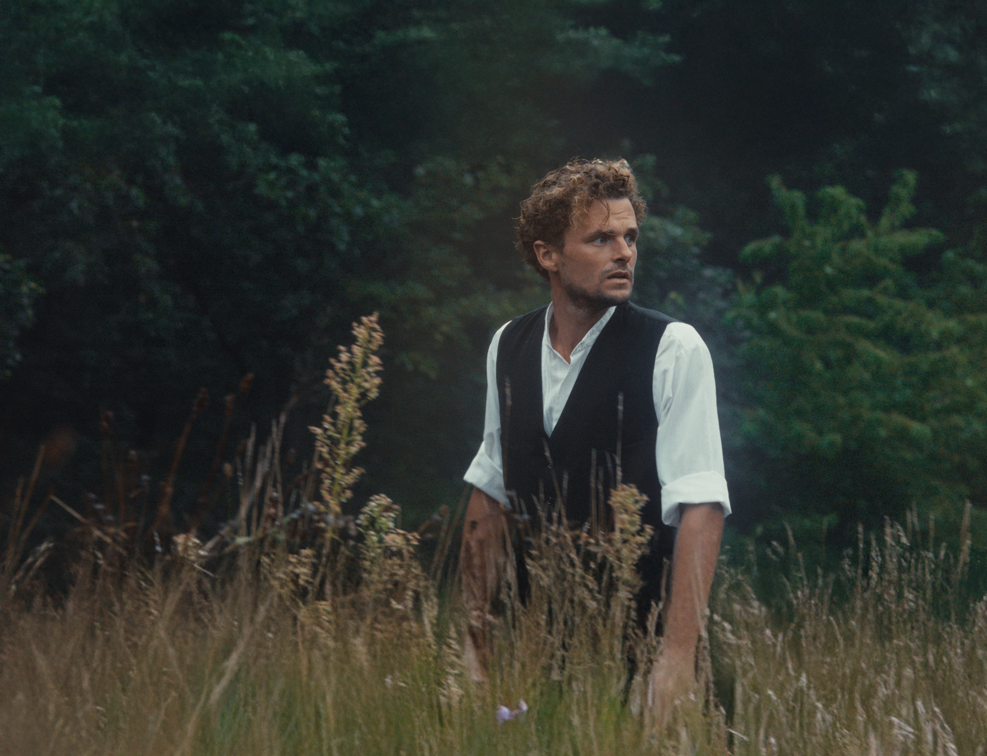 Een persoon met lichtbruin haar, gekleed in een wit shirt en een donker vest, staat in een hoog grasveld en kijkt opzij.