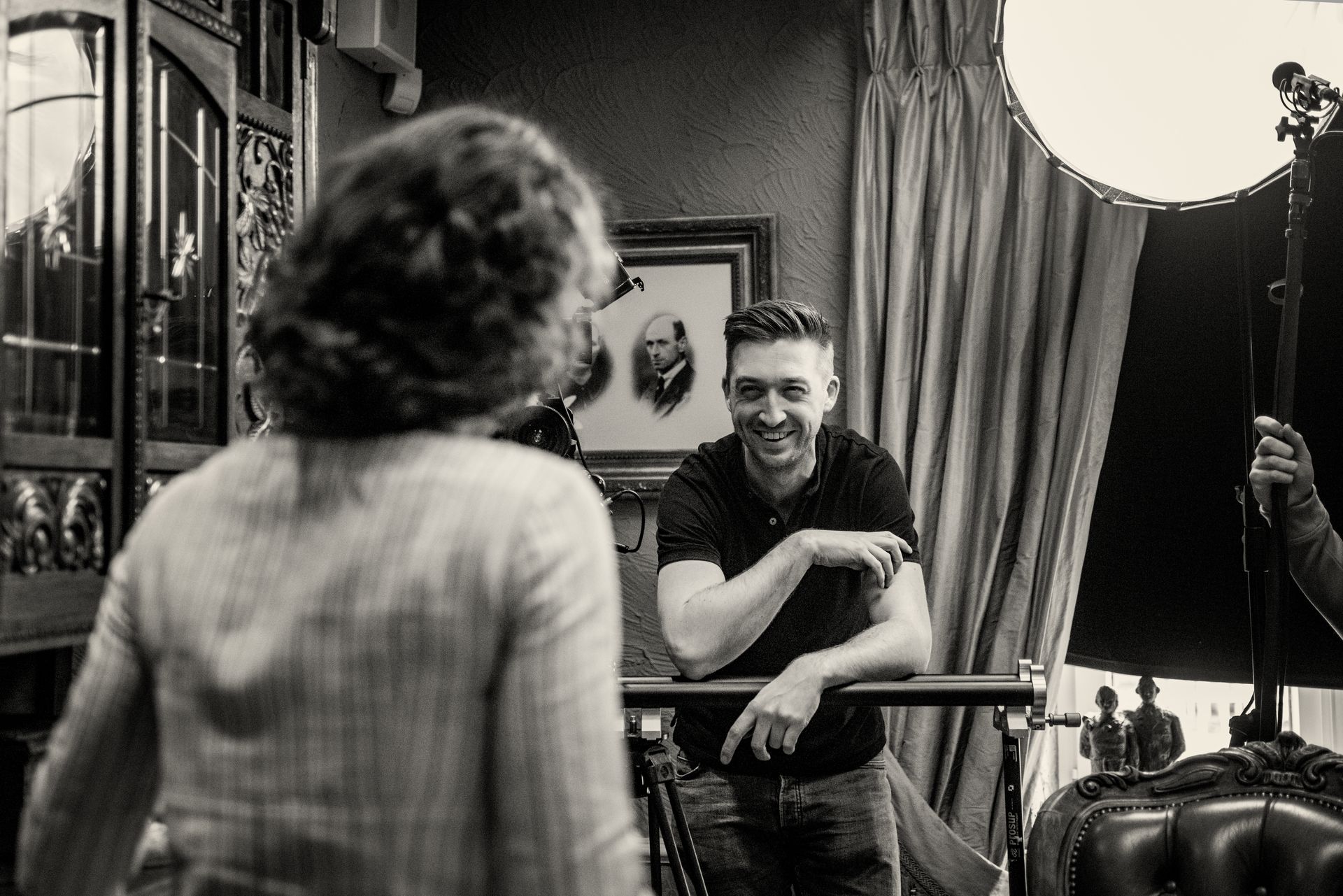 Een lachende man in een donker poloshirt buigt zich over een camerastatief en kijkt naar een persoon met krullend haar in een interieur in vintage-stijl.