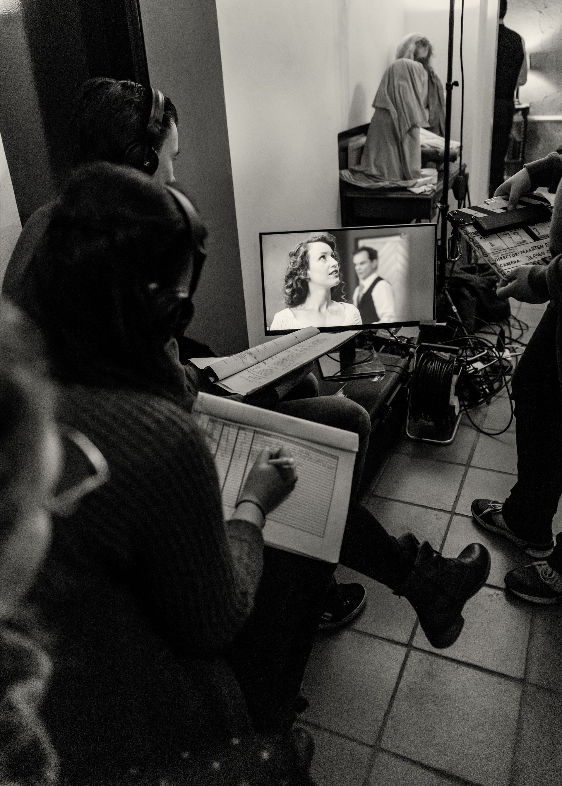 Tijdens het filmen bekijkt een filmploeg een zwart-witscène van een stel op een monitor, waarbij één persoon aantekeningen maakt.