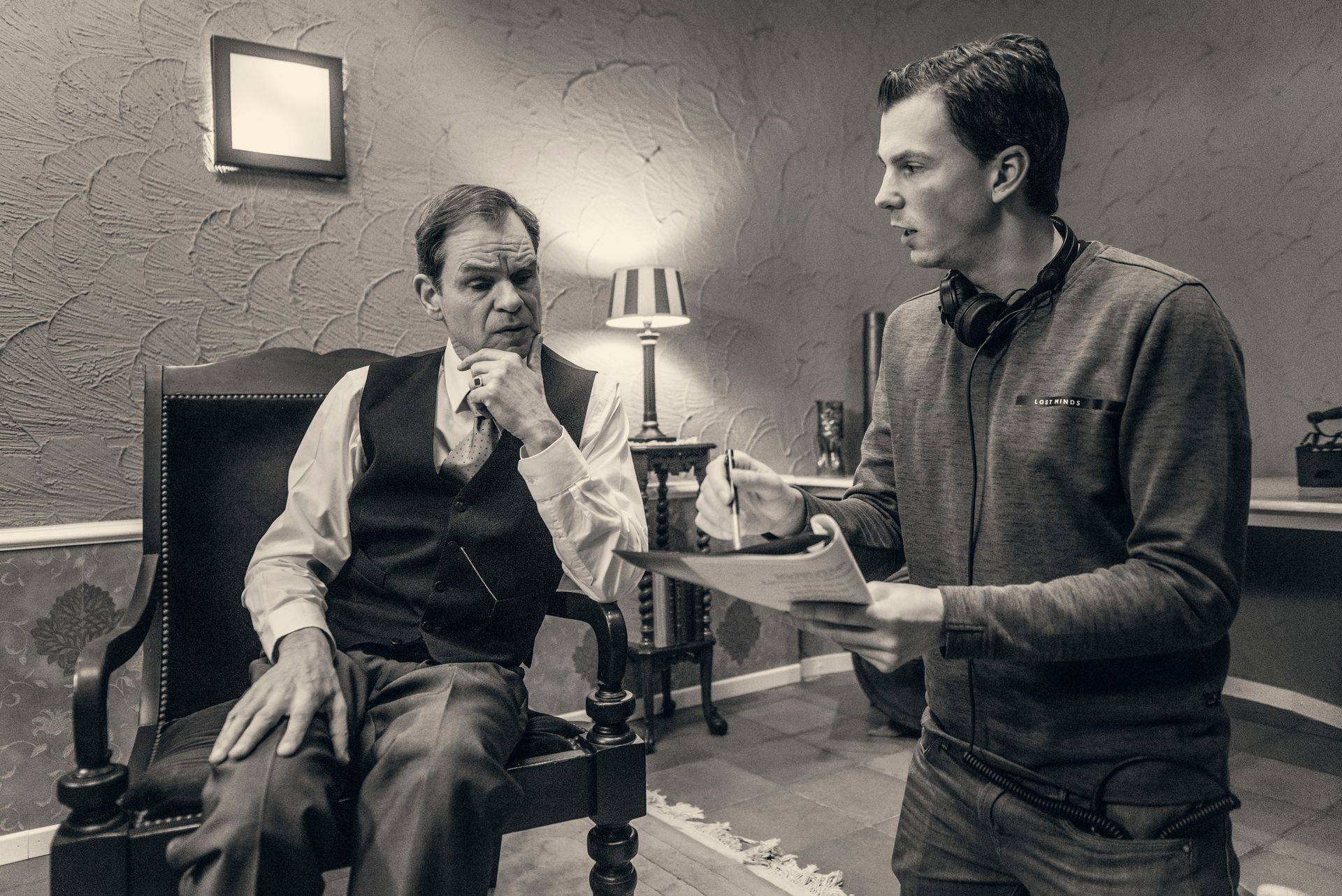 Iemand zit op een stoel terwijl een andere persoon die vlakbij staat een document vasthoudt, kennelijk om instructies te geven of werk te beoordelen.