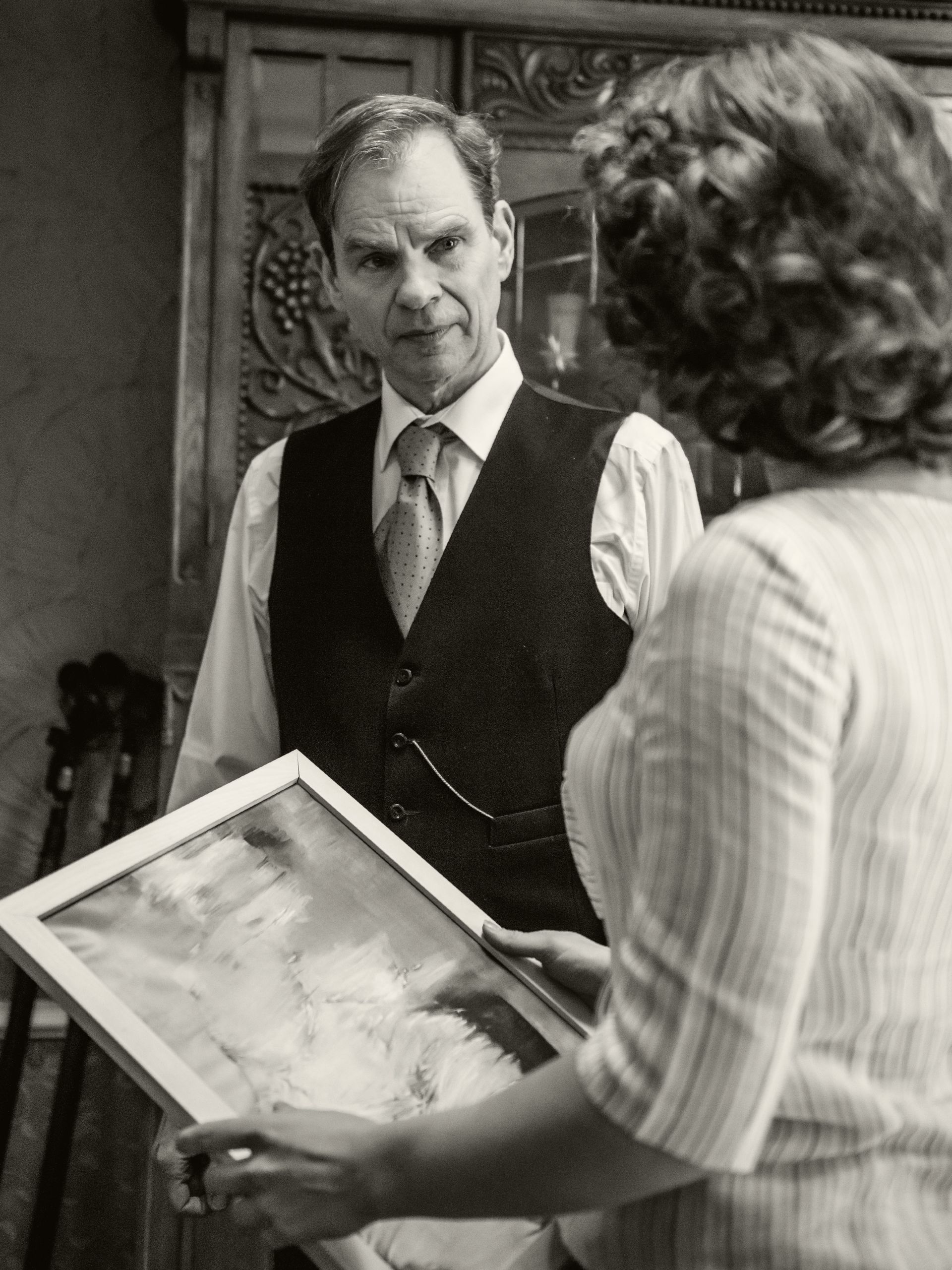 Een persoon in een vest en stropdas kijkt naar iemand anders die een ingelijst kunstwerk vasthoudt in een kamer met houten lambrisering.