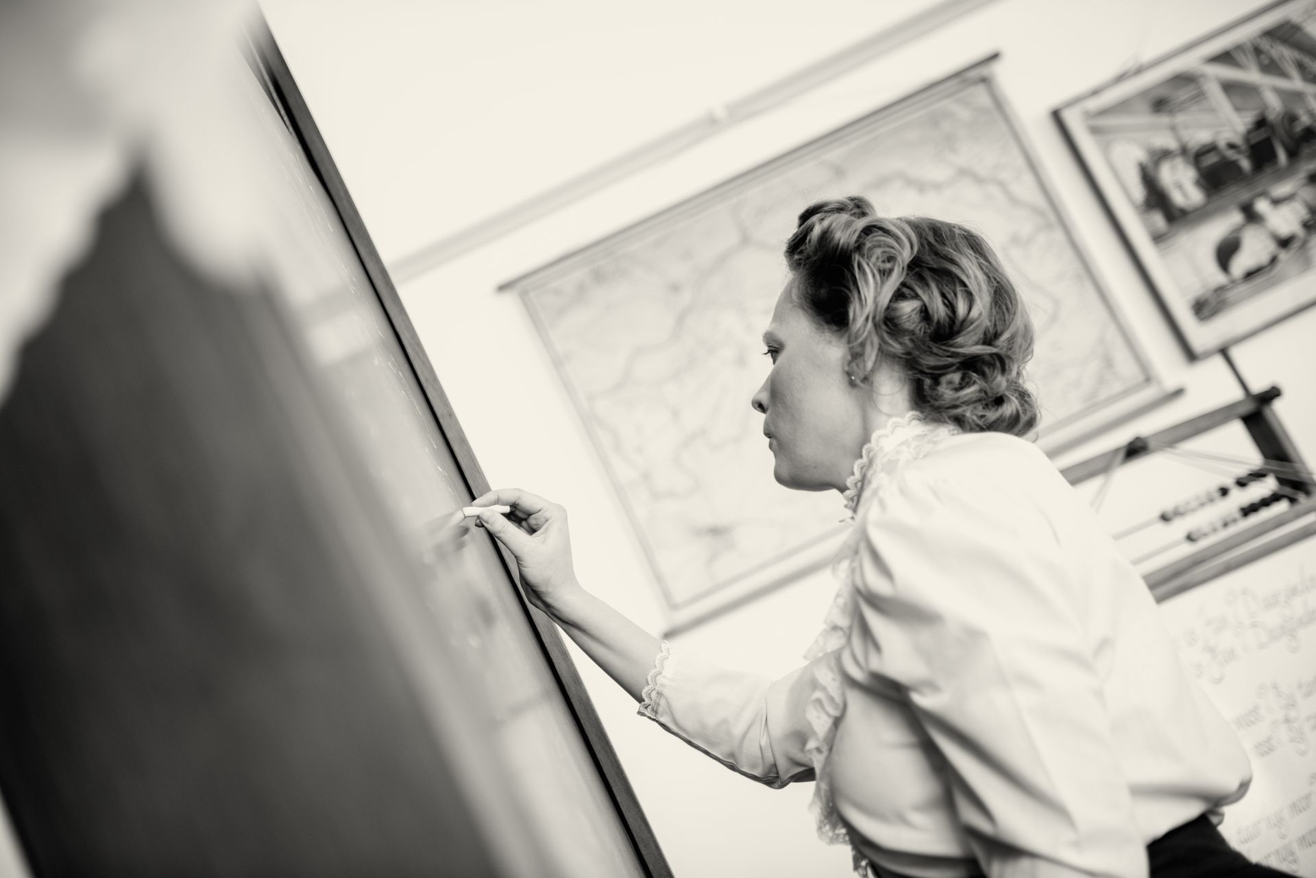 Een persoon in vintage-stijl schrijft op een schoolbord in een kamer met een ingelijste kaart en een telraam aan de muur.