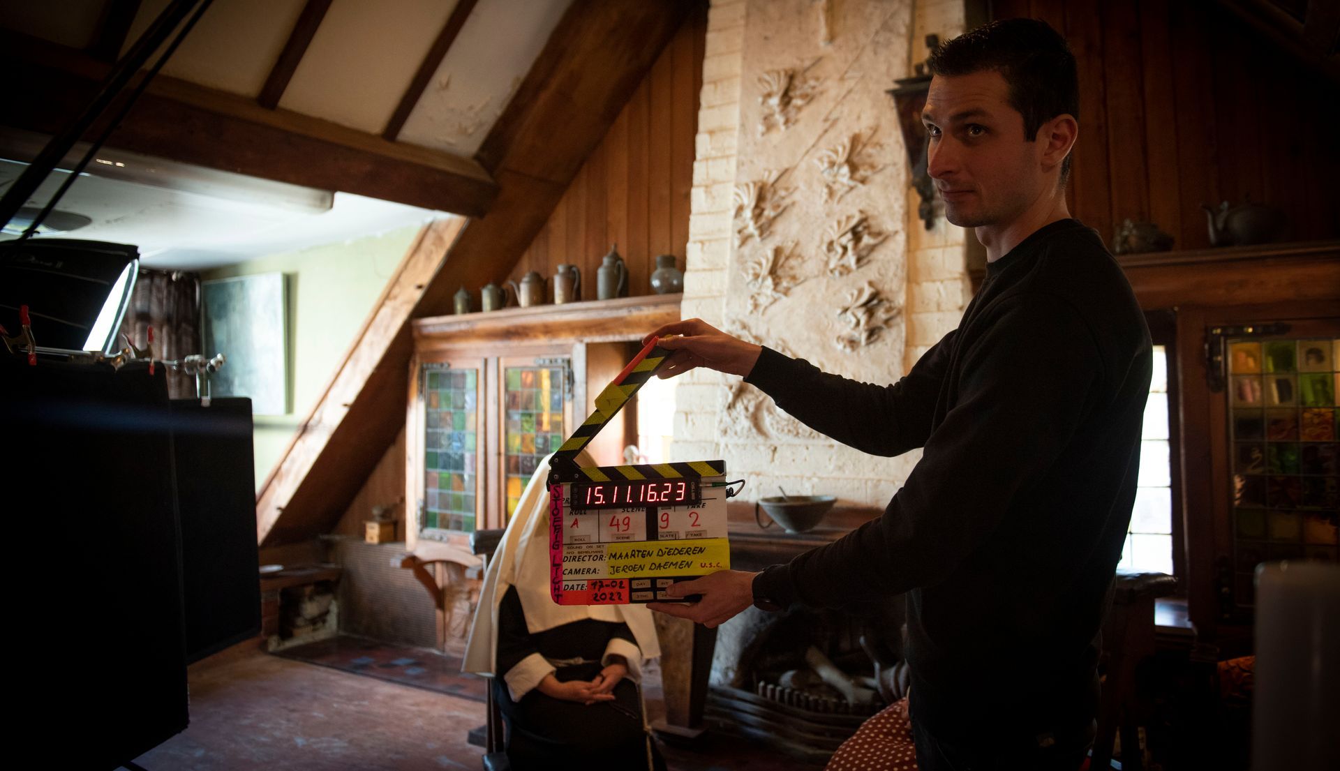 Een persoon houdt een filmklapper vast voor een cameraploeg op een rustieke binnenlocatie met een stenen open haard.