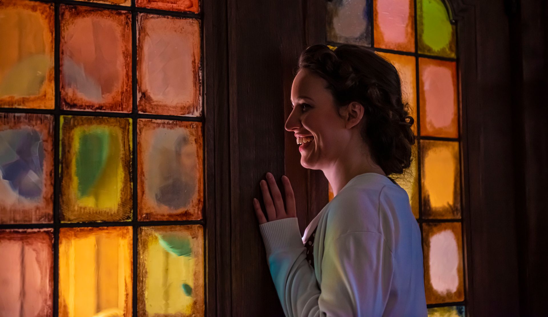 Een glimlachend persoon staat bij een raam met kleurrijke, gebrandschilderde glas-in-loodramen en kijkt naar het licht.