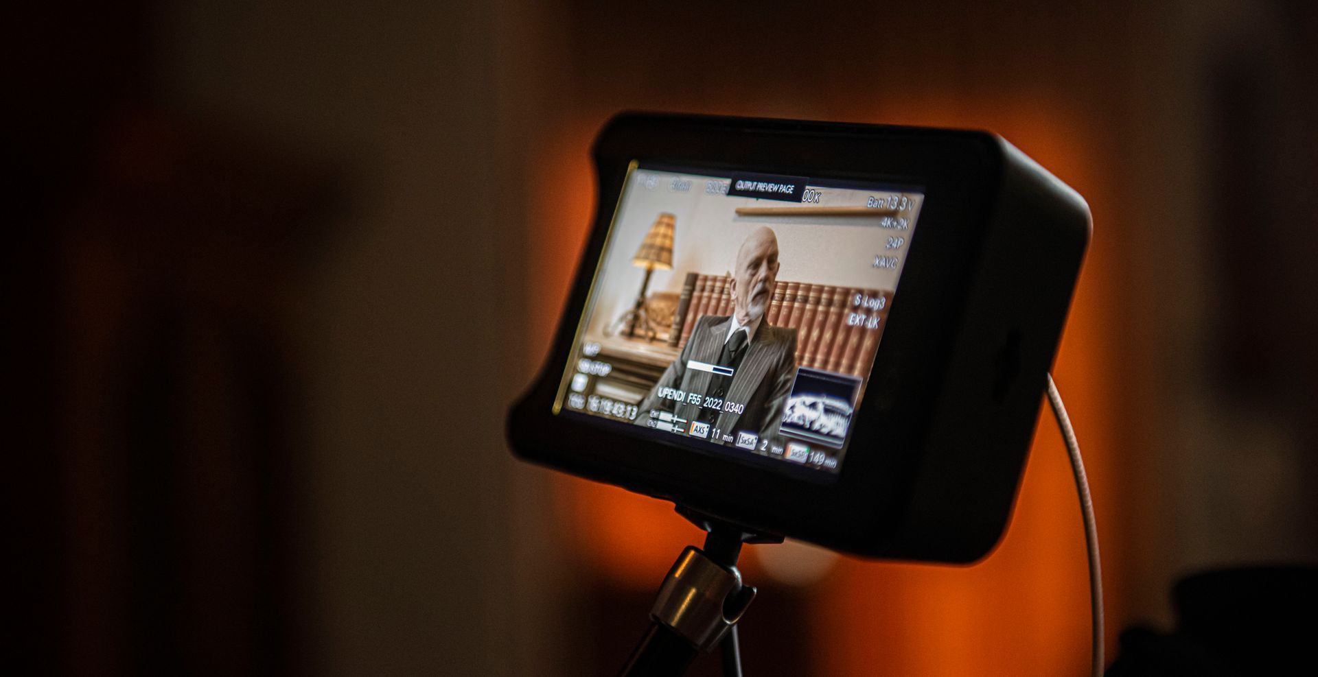 Een cameramonitor op een statief toont een persoon in pak die in een binnenruimte met een lamp zit.