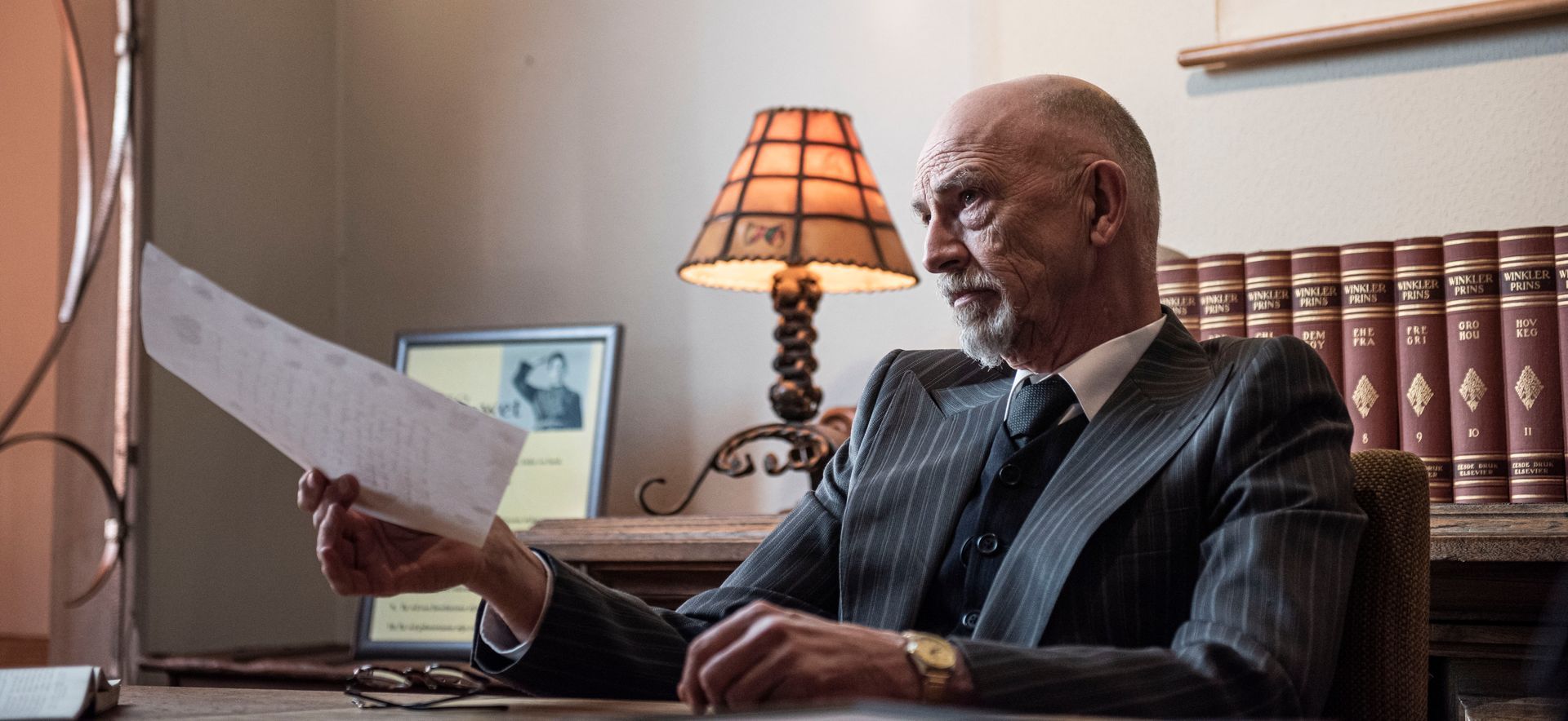 Een persoon in pak zit aan een bureau, houdt een document vast en kijkt opzij in een kamer met een lamp en boekenkasten.