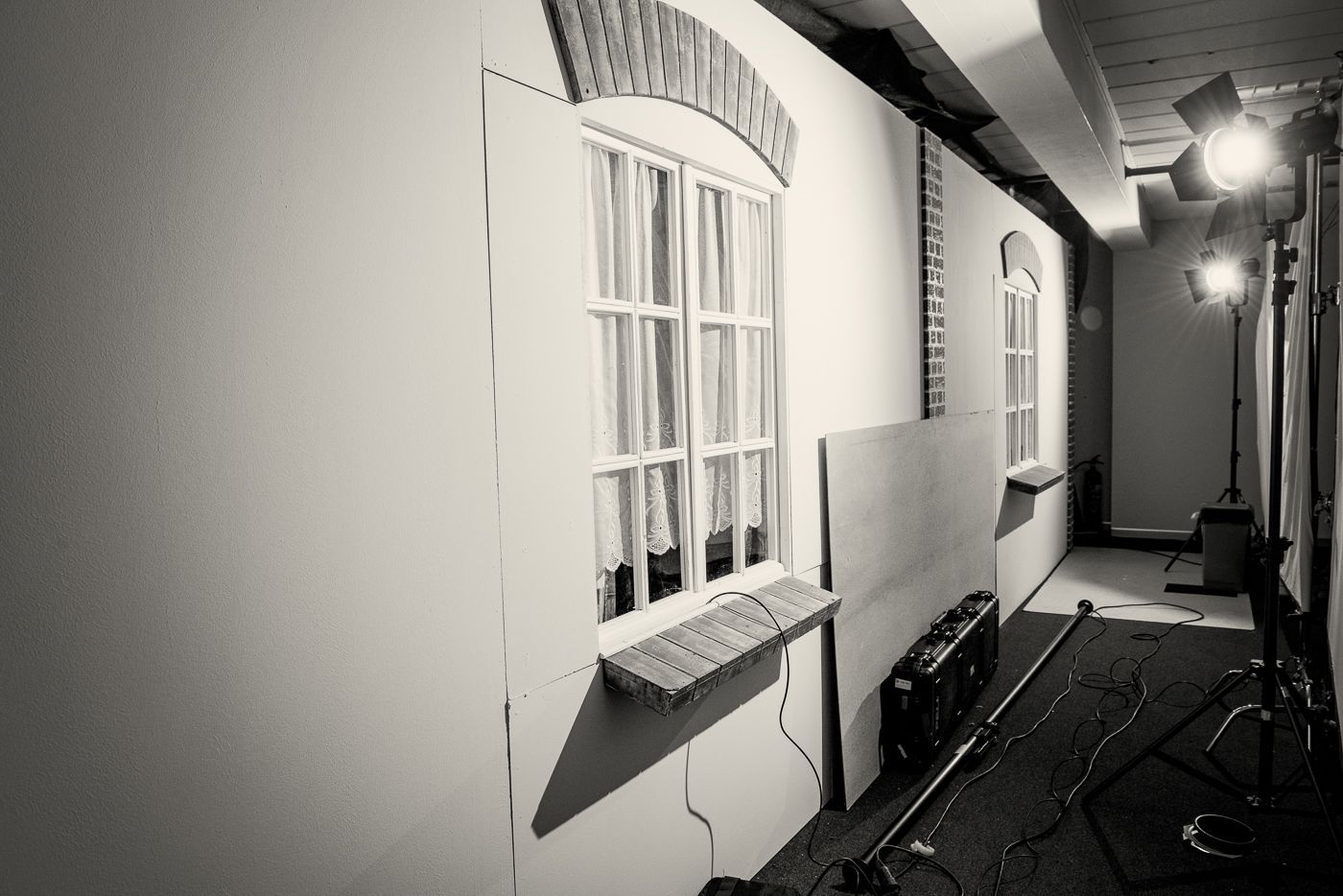 Een filmset met een witte muur met nepramen en bakstenen bogen, verlicht door studiolampen in een smalle ruimte.