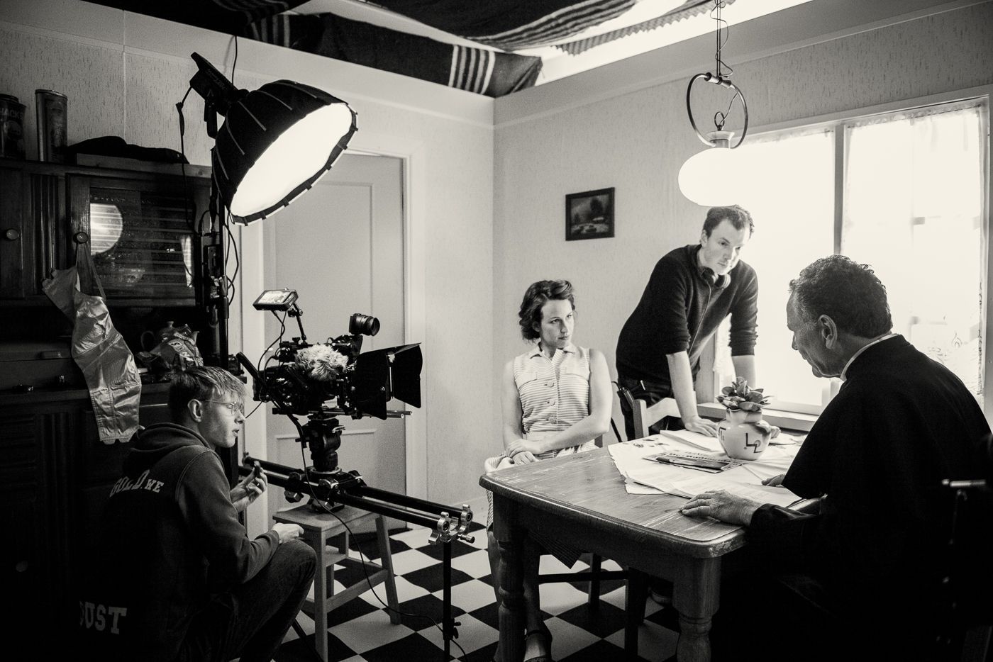 Een filmploeg filmt een scène waarin een persoon aan een tafel zit met een figuur in geestelijke kleding in een zwart-witte ruimte.