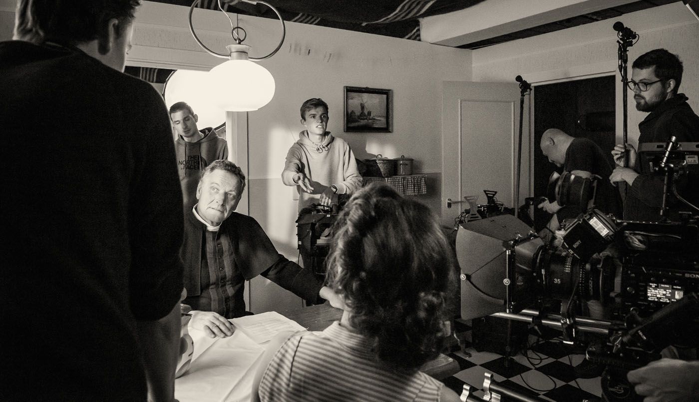 Een zwart-witfoto achter de schermen van een filmploeg die een persoon in geestelijke kleding filmt, zittend aan een tafel.