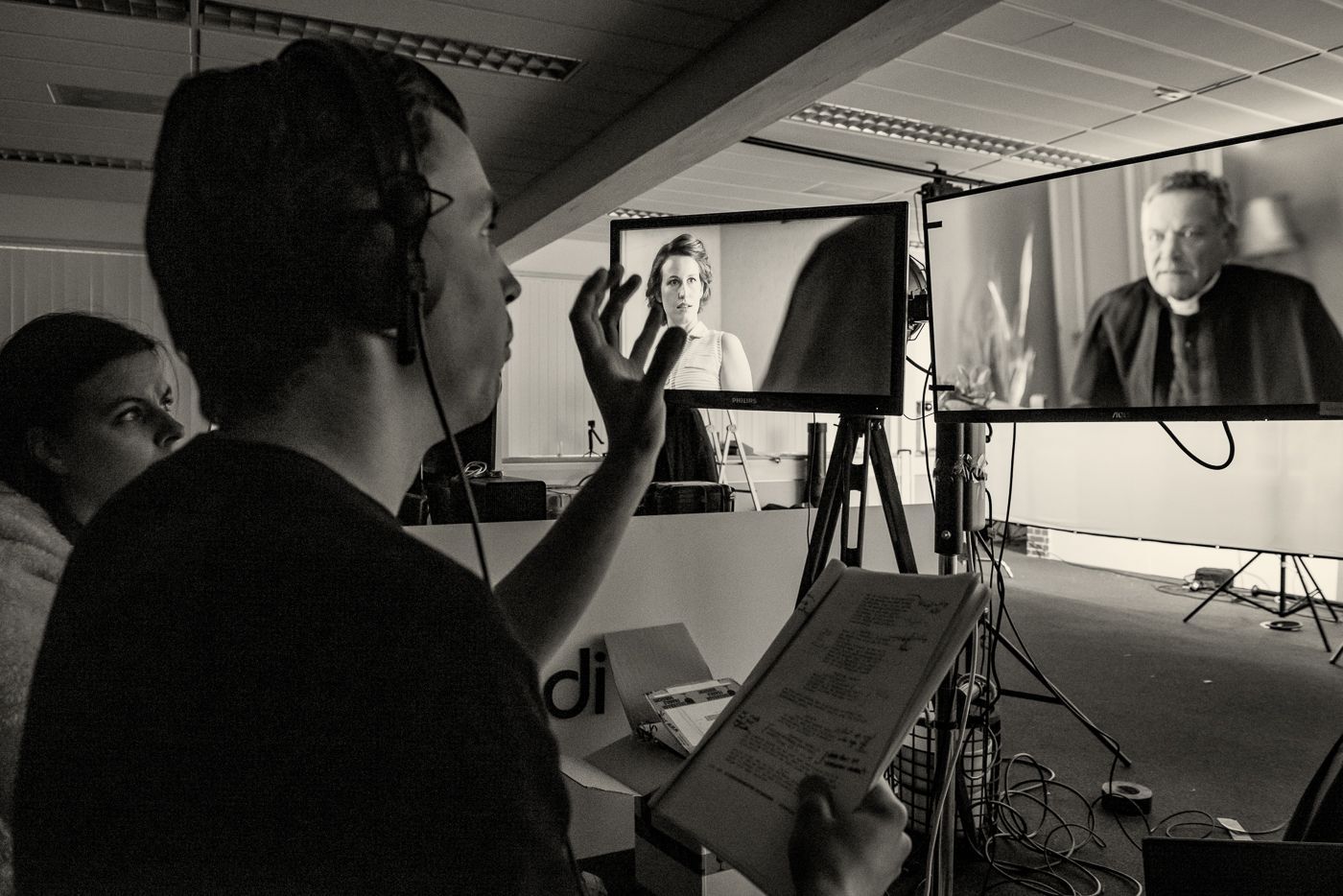 Een persoon met een koptelefoon gebaart naar twee monitoren waarop filmscènes worden getoond, terwijl hij een script vasthoudt in een kantooromgeving.