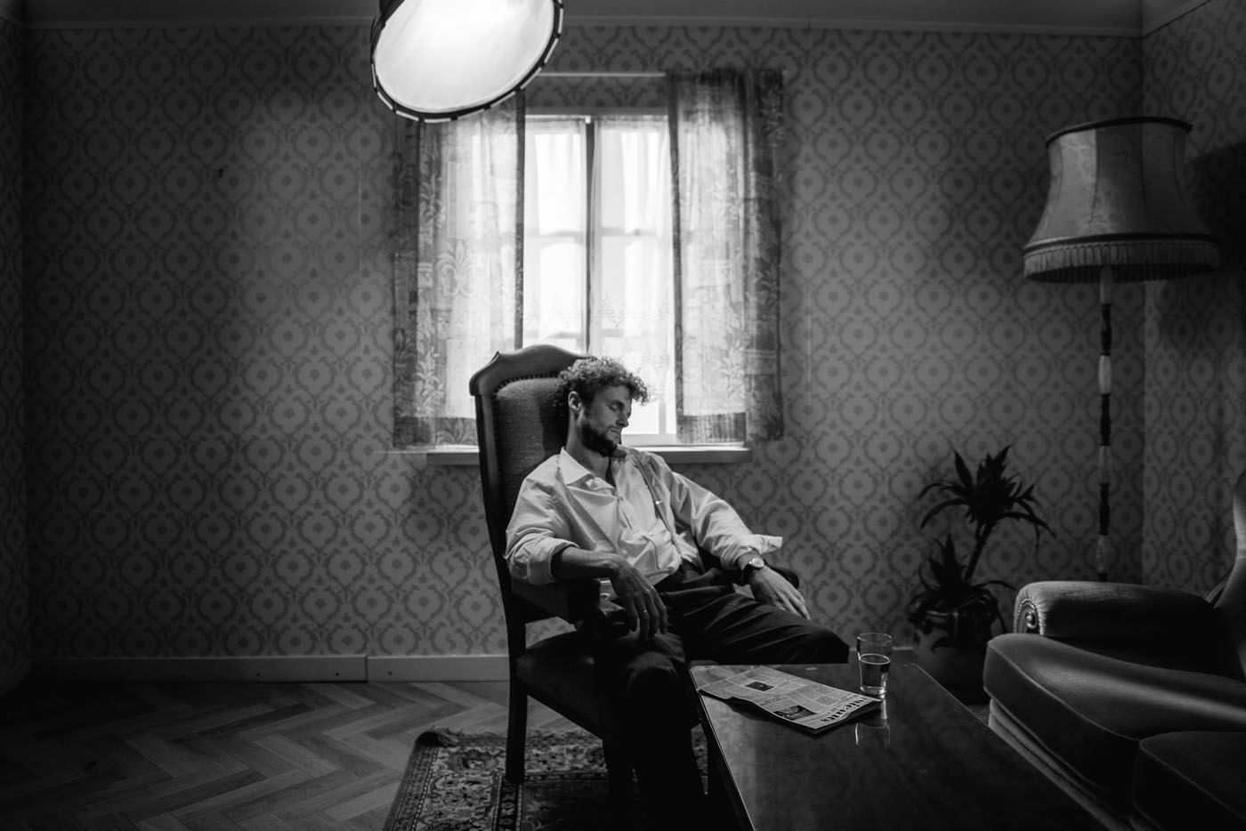 Een persoon zit ontspannen in een fauteuil met hoge rugleuning in een schemerig verlichte kamer in vintage-stijl met behang met een patroon.