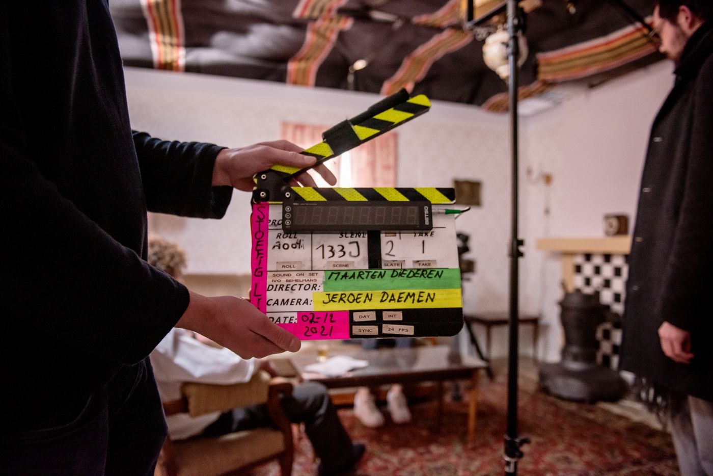 Op de voorgrond van een filmset houdt iemand een filmklapper vast, terwijl op de achtergrond een persoon zit.