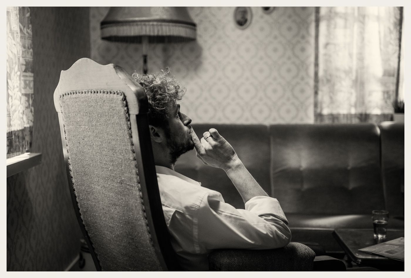 Een persoon met krullend haar zit in een fauteuil, in profielaanzicht, met de kin op de hand rustend in een peinzende houding.