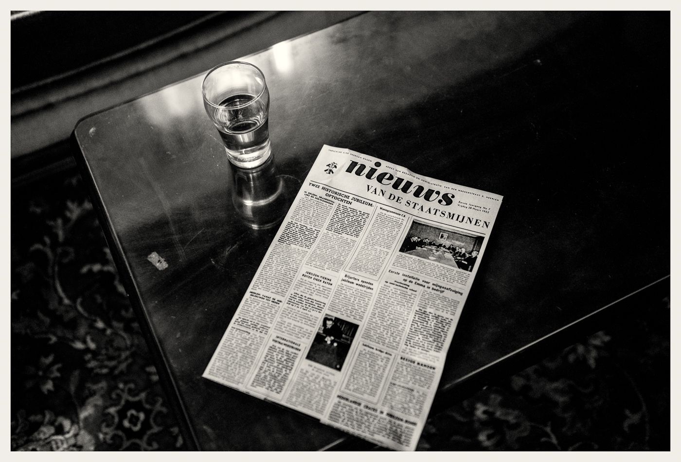 Een krant en een glas water staan op een donkere, reflecterende tafel, vastgelegd in zwart-wit.