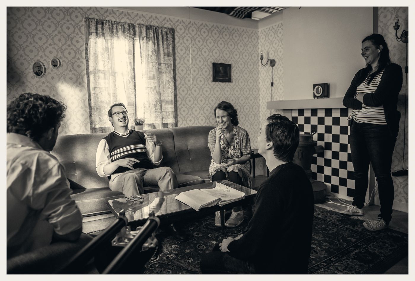 Een groep mensen zit en staat in een woonkamer in vintage-stijl, lachend en pratend rond een salontafel.