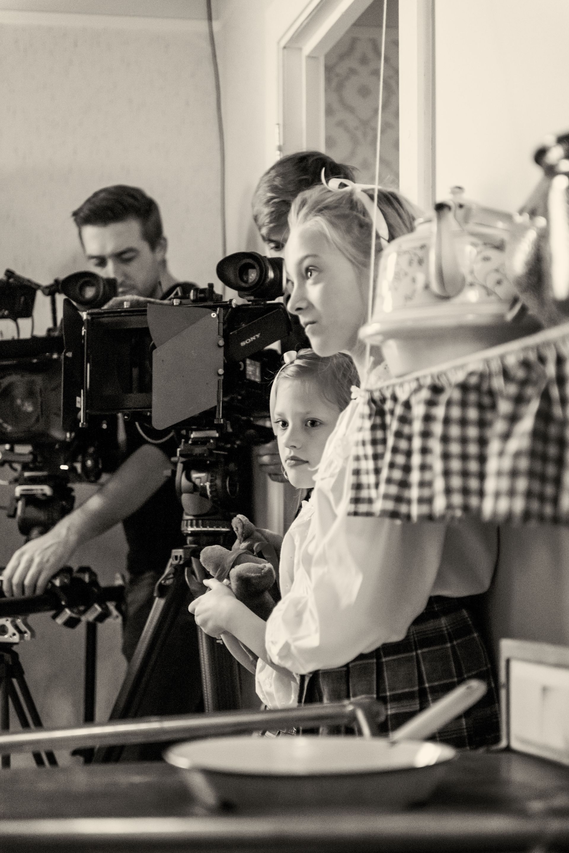 Een persoon bedient een filmcamera op een statief, terwijl twee anderen in de buurt staan in een keukendecor, vastgelegd in sepiatinten.