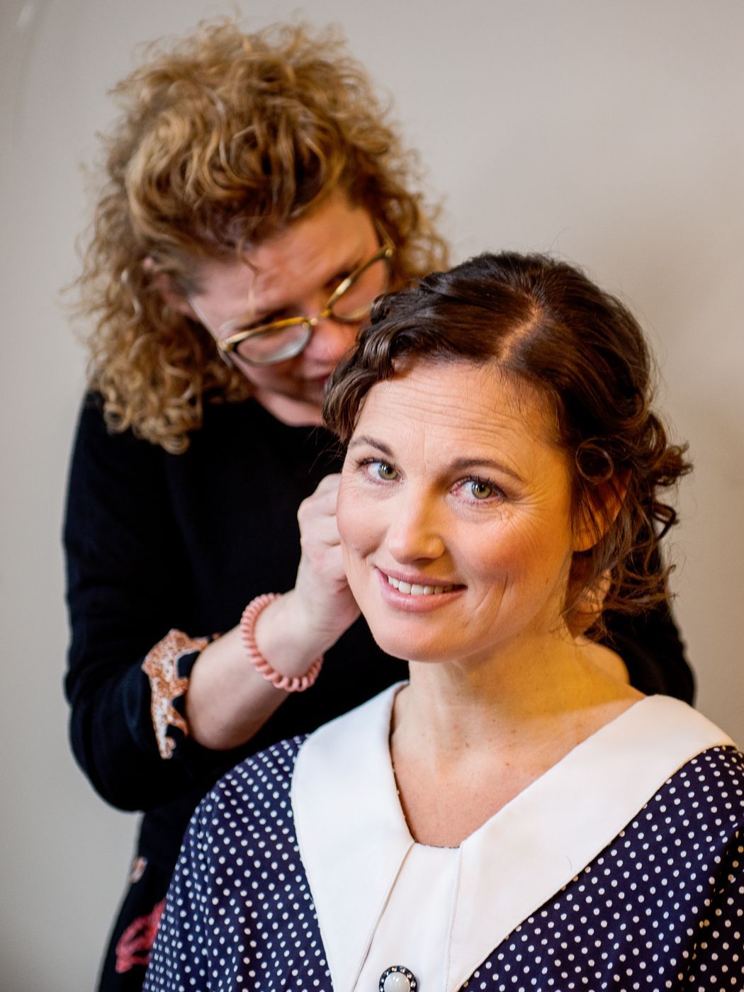 Een styliste helpt een lachende vrouw in een blauwe jurk met stippen oorbellen in te doen.