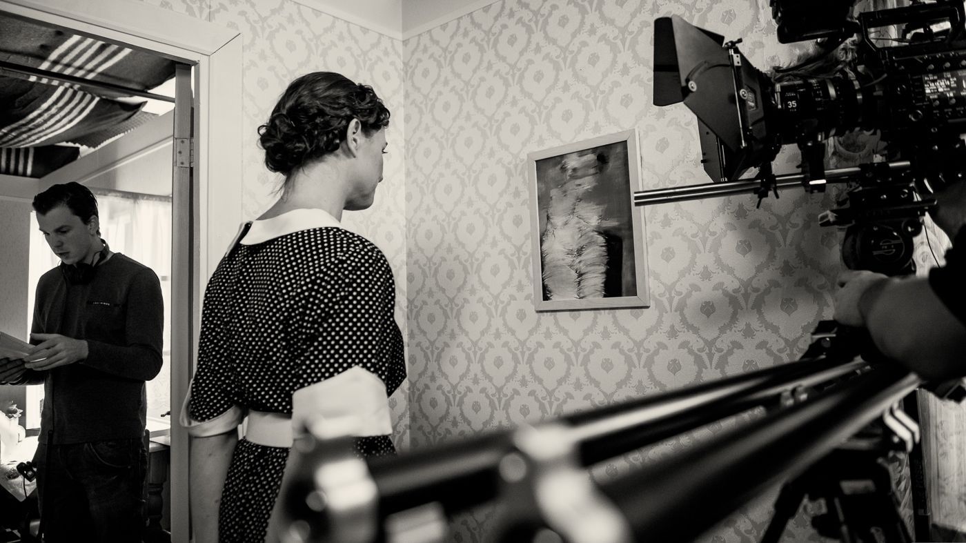 Een filmploeg filmt een scène waarin een persoon in een jurk met stippen voor een ingelijst kunstwerk staat aan een muur met een patroon.