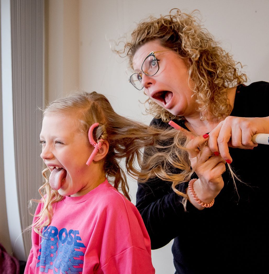 Een persoon met krullend haar stylet het haar van een jong kind met een krultang; beiden kijken verbaasd en hebben hun mond open.
