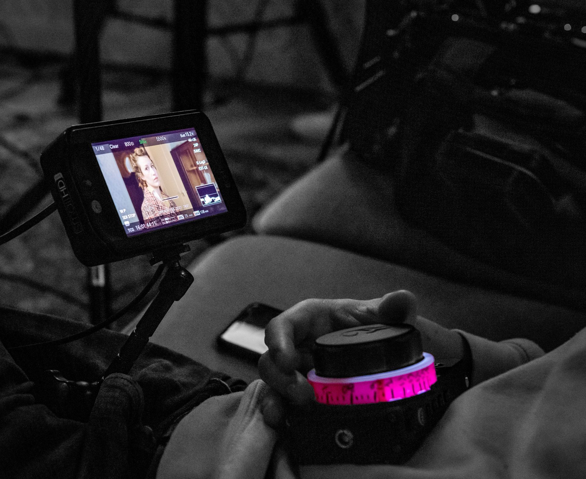 Een persoon houdt een draadloze scherpstelcontroller met een oplichtende roze ring vast terwijl hij een videobeeld op een klein extern scherm bekijkt.