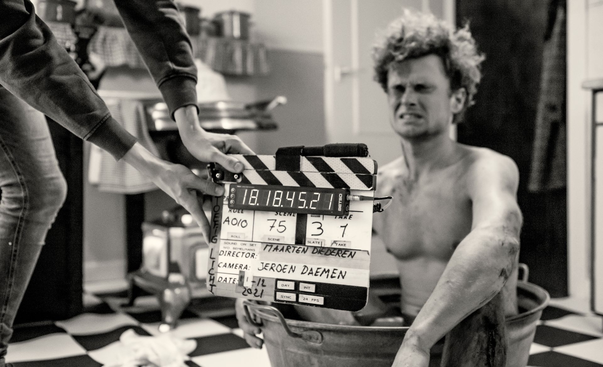 Een filmklapper wordt voor een persoon met ontbloot bovenlijf gehouden, die in een metalen bak zit in een zwart-wit setting achter de schermen.