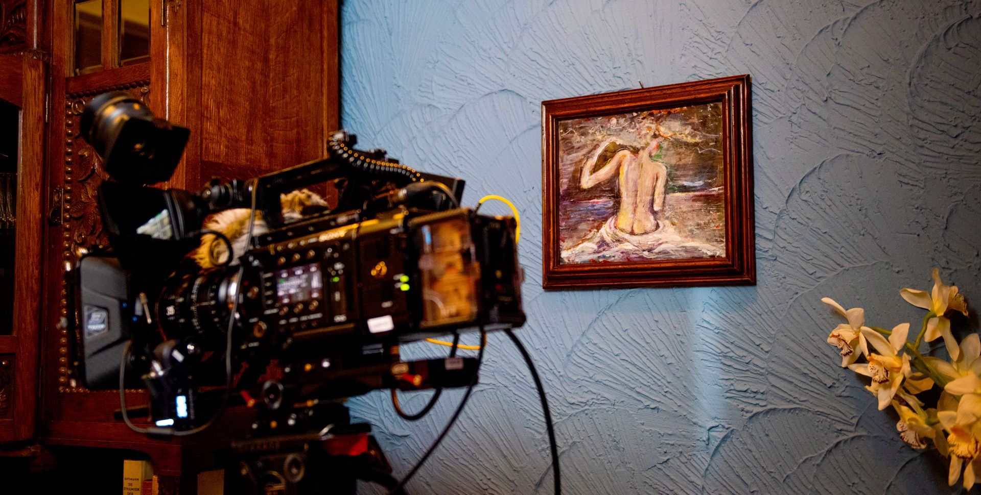 Een professionele filmcamera op een statief is gericht op een blauwe muur met structuur, waarop een ingelijst impressionistisch schilderij en bloemen te zien zijn.