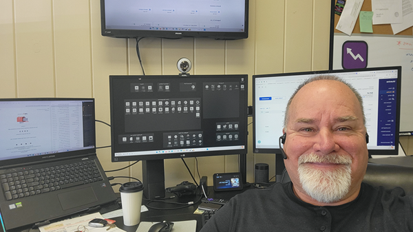 Man with headset smiles at the camera, seated at desk with multiple monitors, laptop, and coffee.