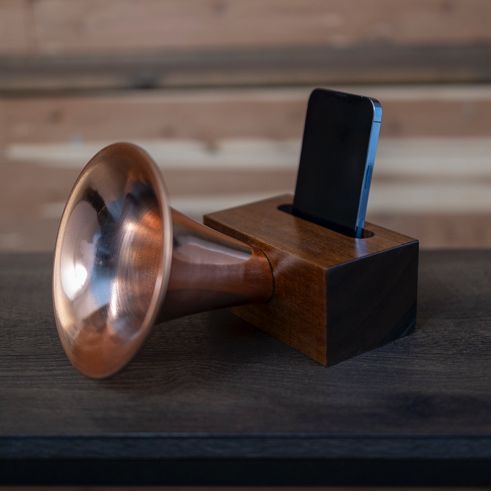 A copper trumpet sits on a wooden table next to a cell phone