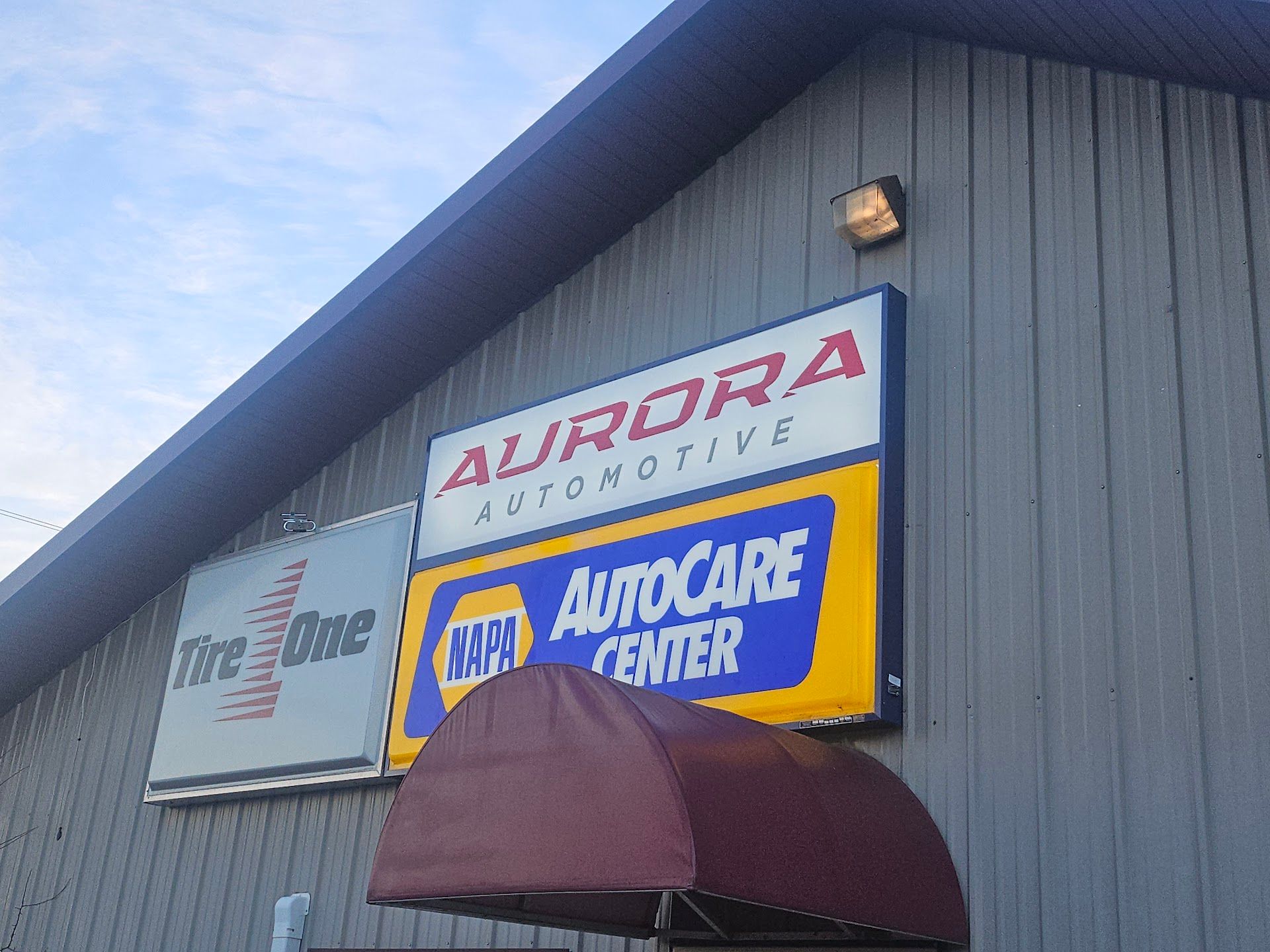 A gray metal building with signs for Aurora Automotive, Tire One, and a NAPA AutoCare Center under a maroon awning. | Aurora Automotive