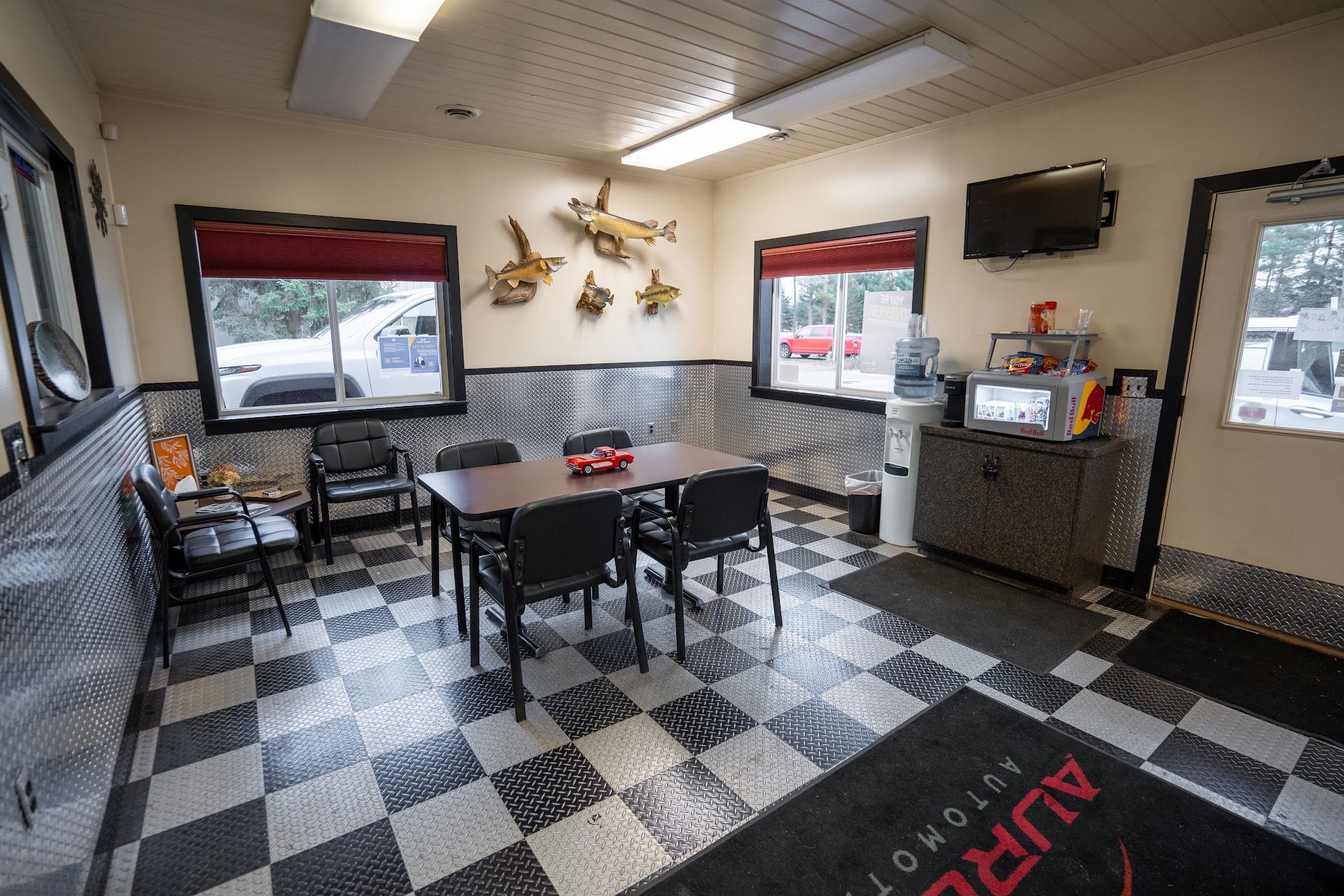 A waiting area with a checkered floor, a central table and chairs, wall-mounted fish decor, and a television. | Aurora Automotive