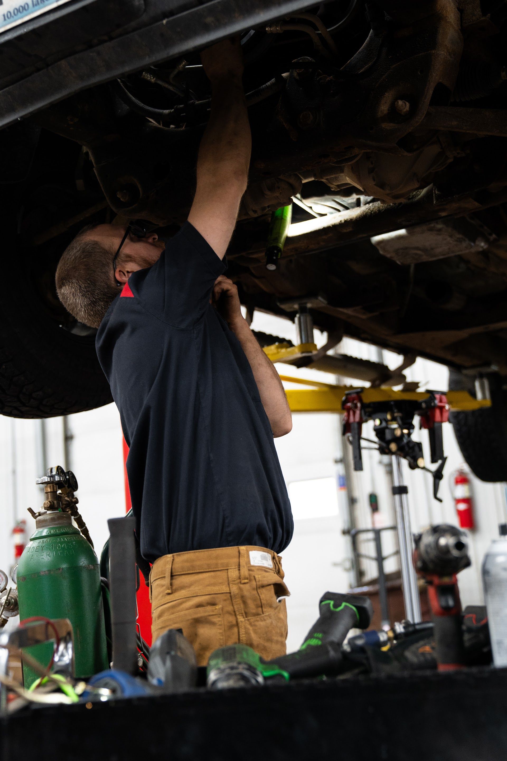A mechanic wearing a dark shirt and tan pants works on the undercarriage of a raised vehicle in a repair shop. | Aurora Automotive