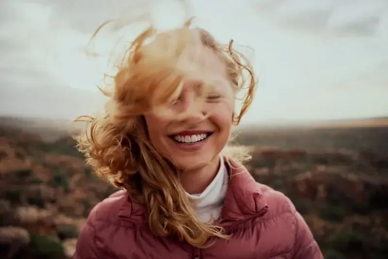 Kvinne som smiler, med vind i håret, utendørs, iført rosa jakke, naturskjønn bakgrunn.