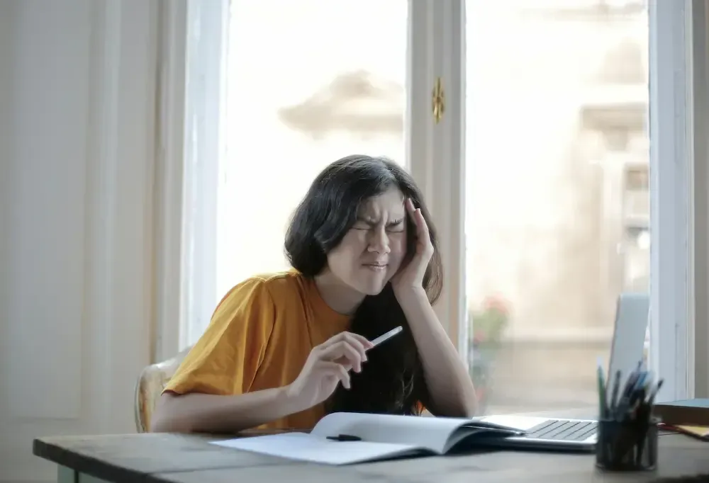 Kvinne ved skrivebordet, hånden på hodet, øynene knyttet sammen, ser stresset ut mens hun jobber på bærbar PC og notatbok.