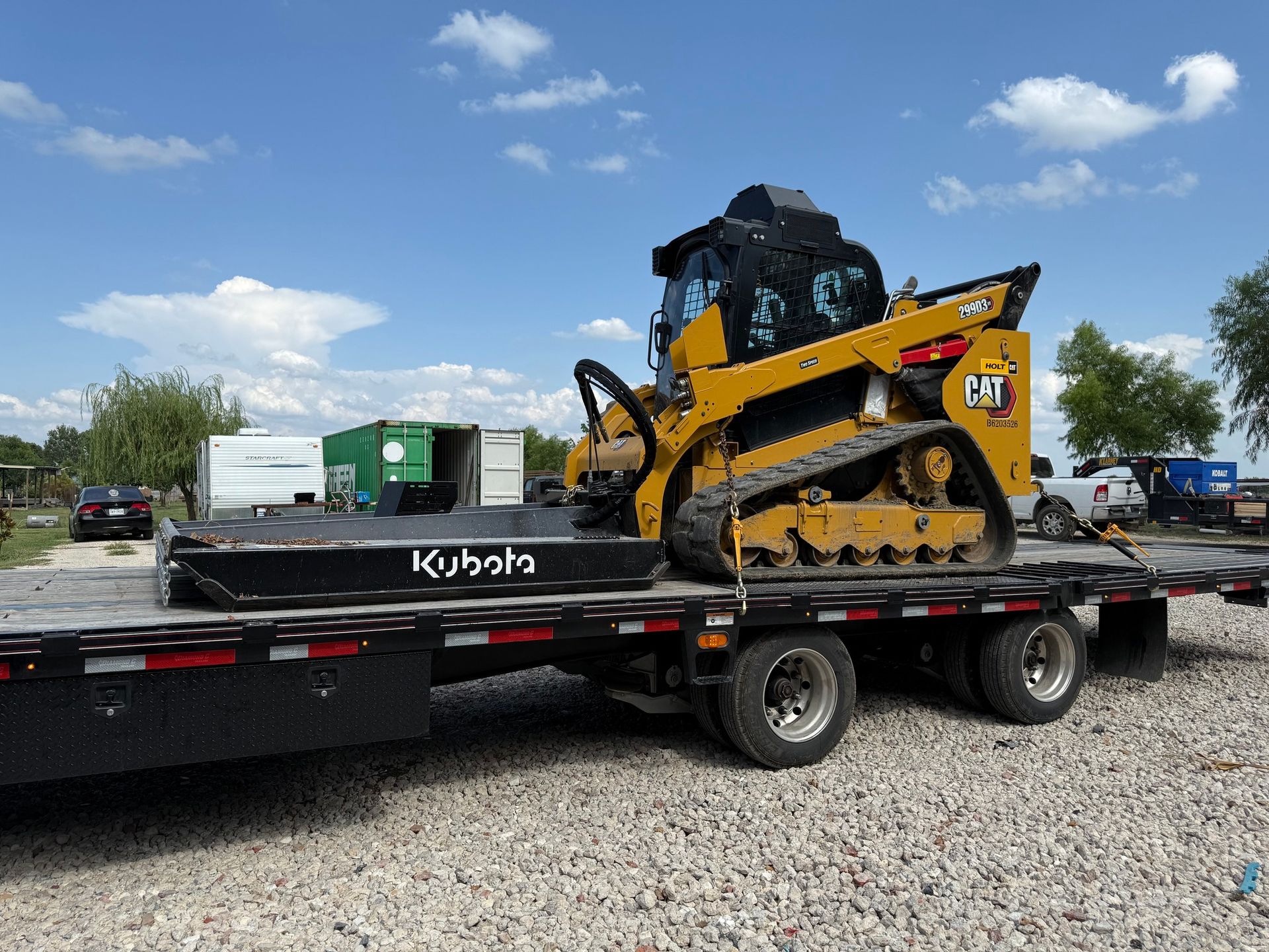 Yellow Caterpillar track loader and black Kubota attachment on a trailer, outdoors.