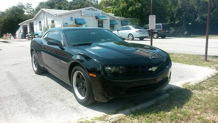 A black Chevrolet Camaro parked in a gravel lot in front of a white building on a sunny day.