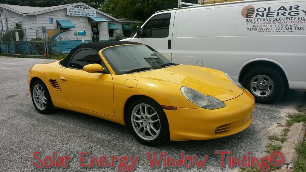 A bright yellow Porsche convertible parked in a gravel lot next to a white service van.