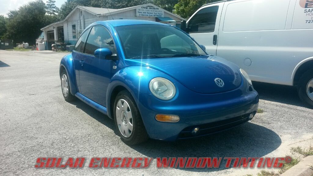 A shiny blue Volkswagen Beetle parked on a gravel lot near a building and a white work van.