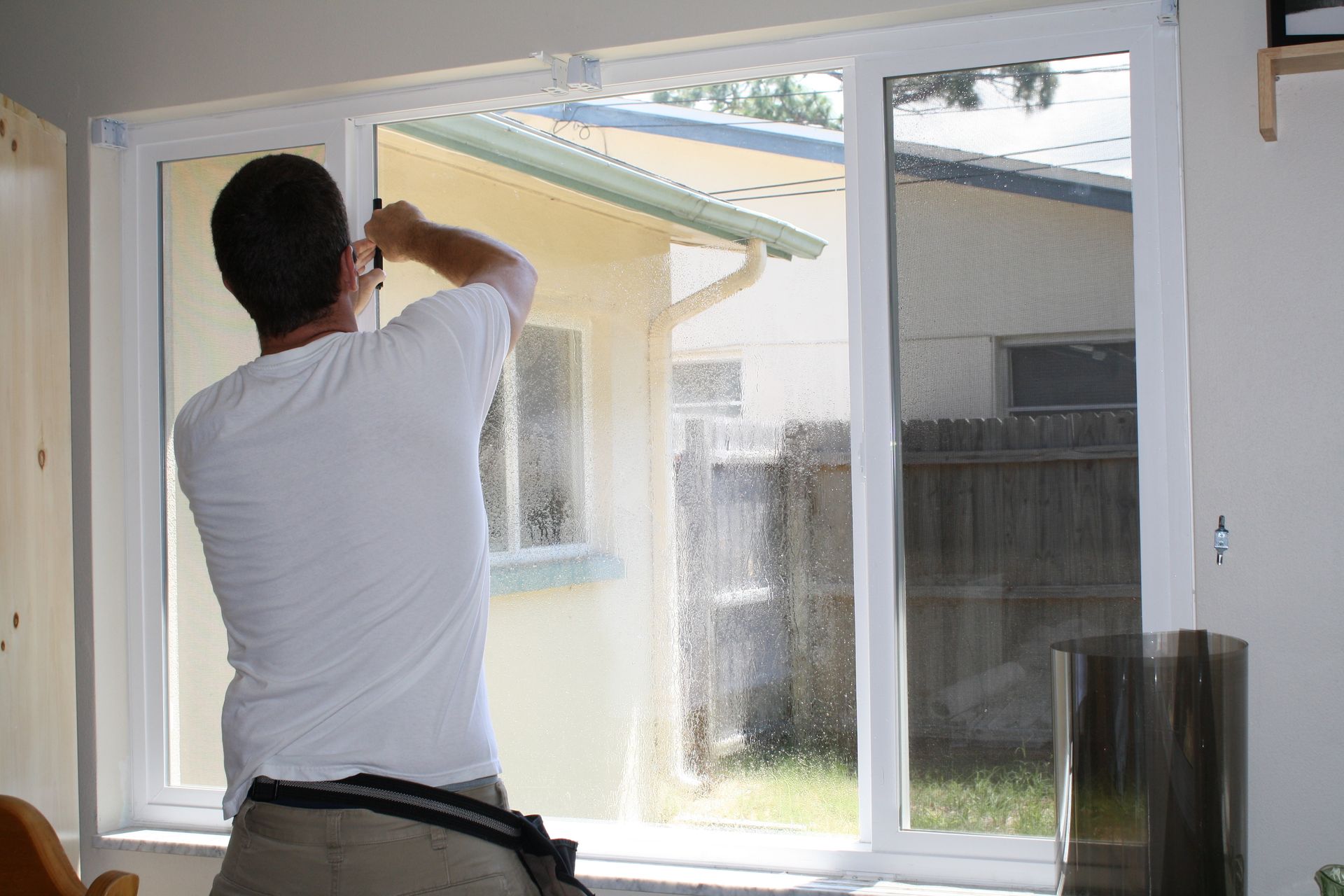 A person in a white shirt installs a window screen inside a room, viewed from behind.