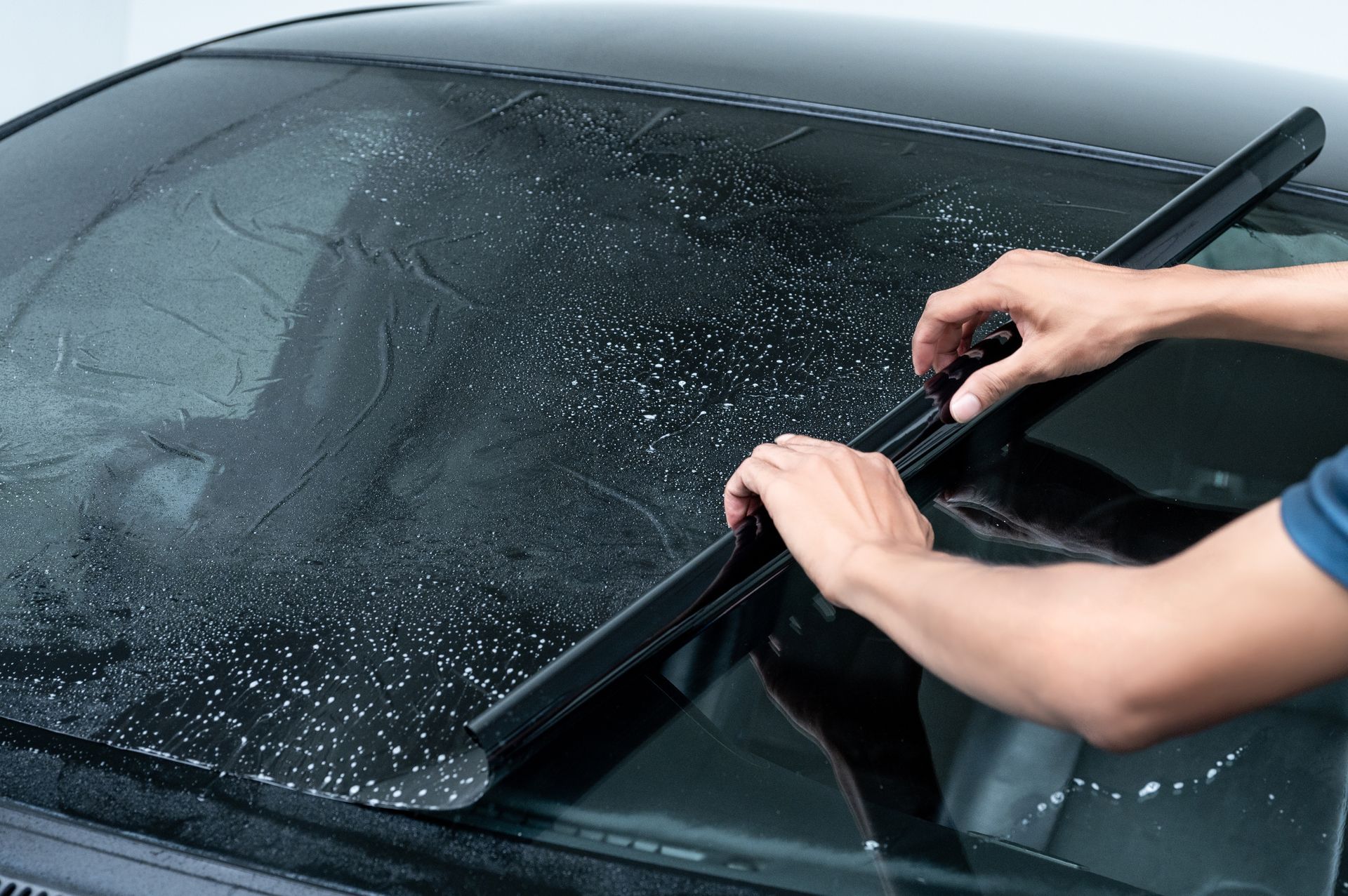 A person uses a squeegee to smooth out a sheet of dark window tint film onto the wet glass of a car windshield.