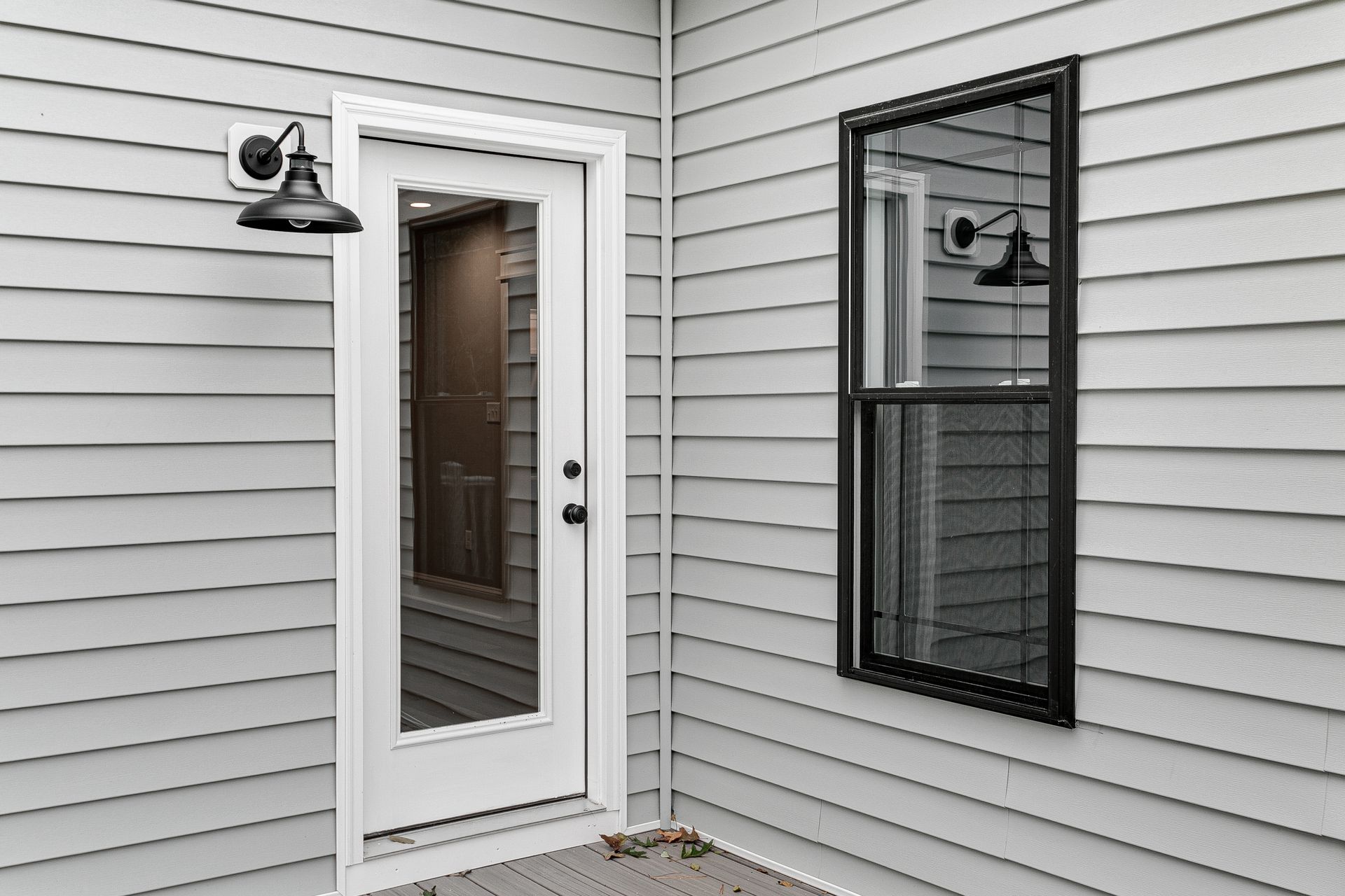A white door and a tall black-framed window on a grey-sided exterior wall, illuminated by a black wall sconce.