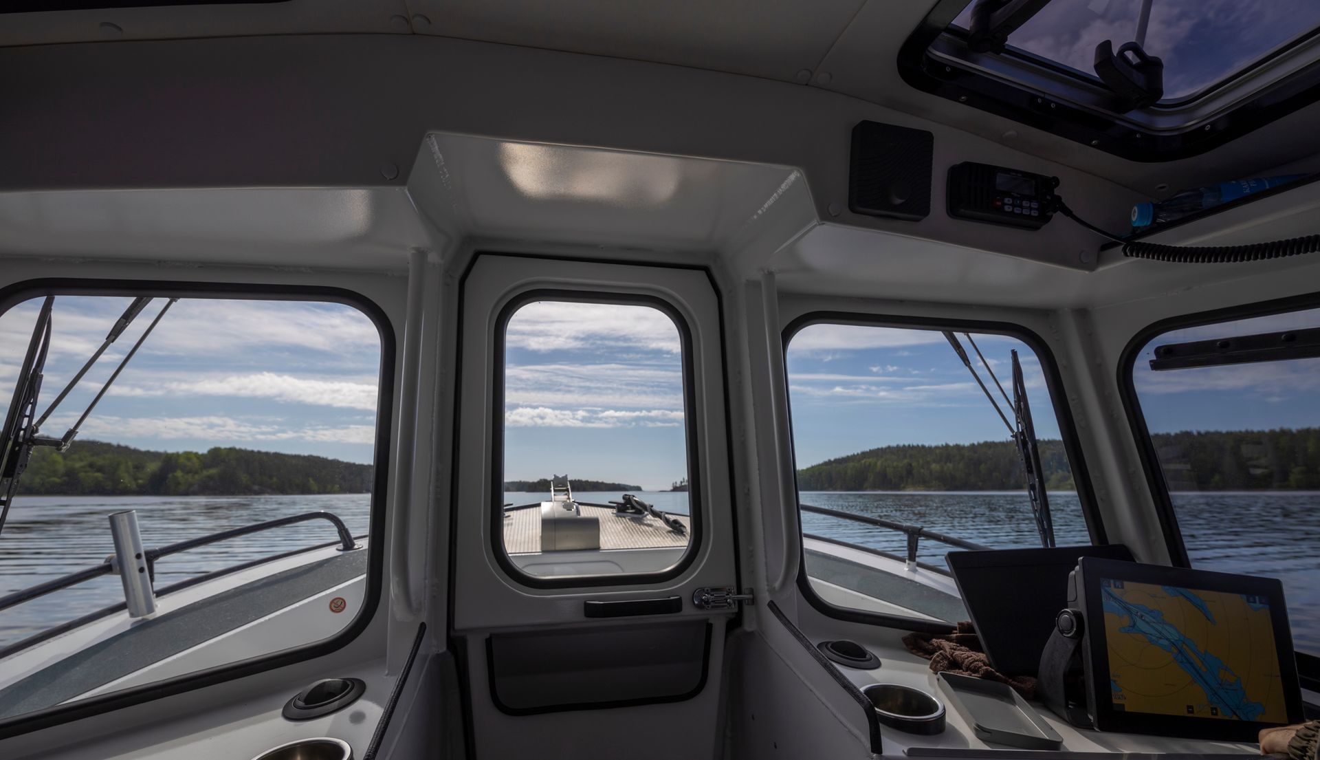 View from inside a boat cabin looking out through windows at a calm lake surrounded by forested shores.