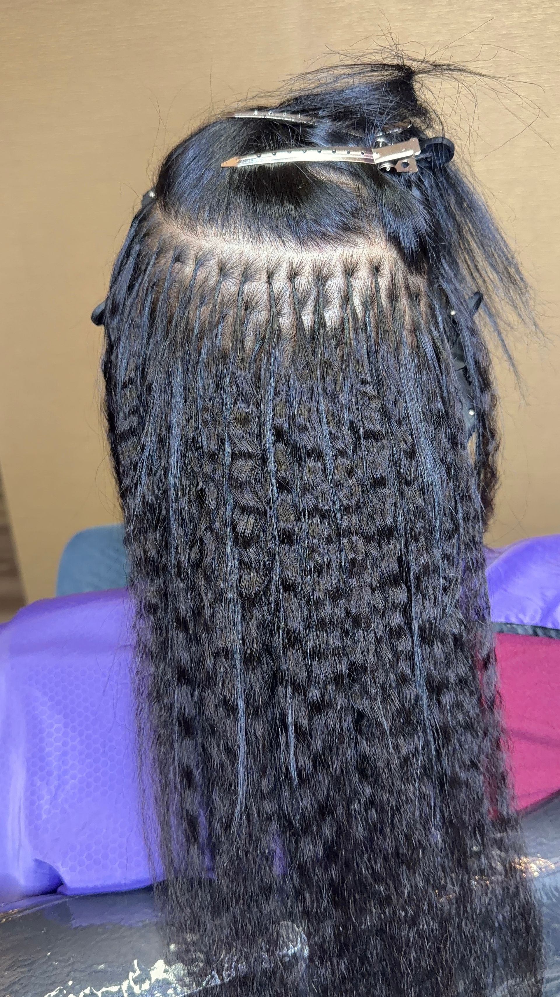 A back view of a person receiving micro-link hair extensions, showing dark, curly hair attached to small base sections.