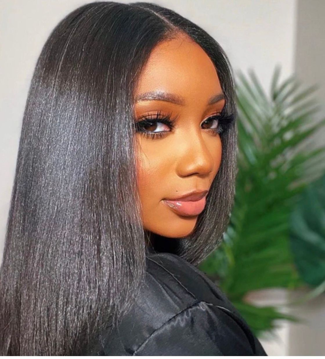 A person with sleek, dark hair looks over their shoulder at the camera, wearing neutral makeup in a softly lit setting.