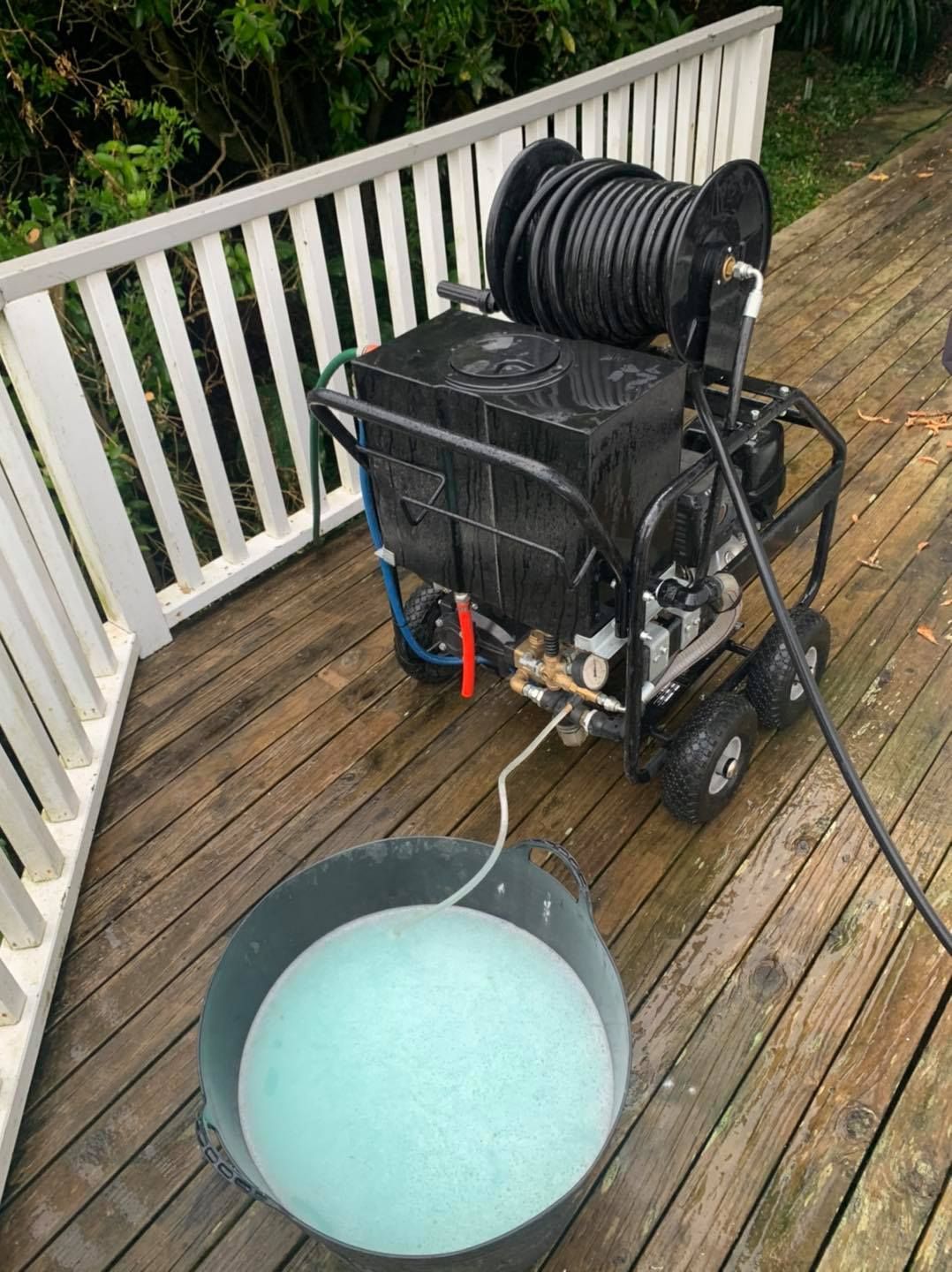 A pressure washer is sitting on a wooden deck next to a bucket of liquid.
