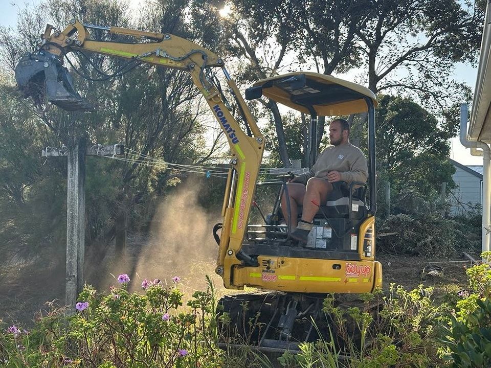 A man is driving a small yellow excavator in a yard.